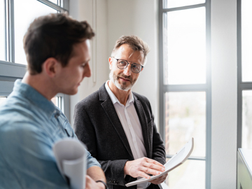 Auf dem Bild sind ein jüngerer und ein älterer Mann mit Brille zu sehen, die sich unterhalten.